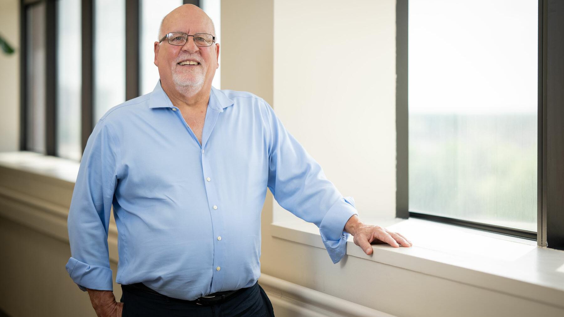 Smiling man in a blue shirt stands near a window