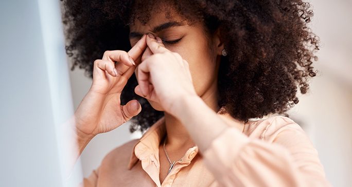Curly-haired woman wearing a peach shirt uses index and middle fingers from both hands to plug her nose.