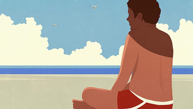 Fair-skinned male with no shirt on sitting on the sand near the beach 