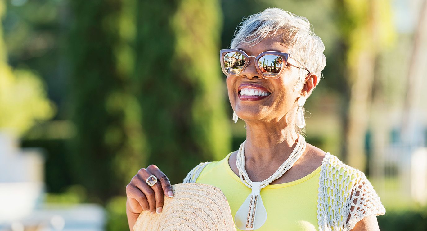 Older woman with darker skin stands smiling in the sun wearing sunglasses and holding a sun hat.