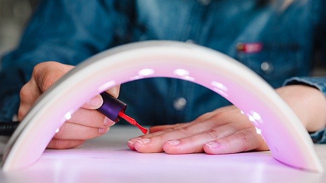 A person paints their nails red behind a white UV light with purple rays.
