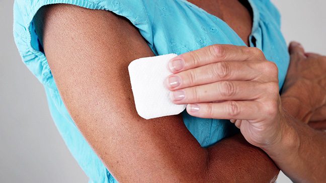 Person in light blue blouse applies a white nicotine patch to their upper arm.