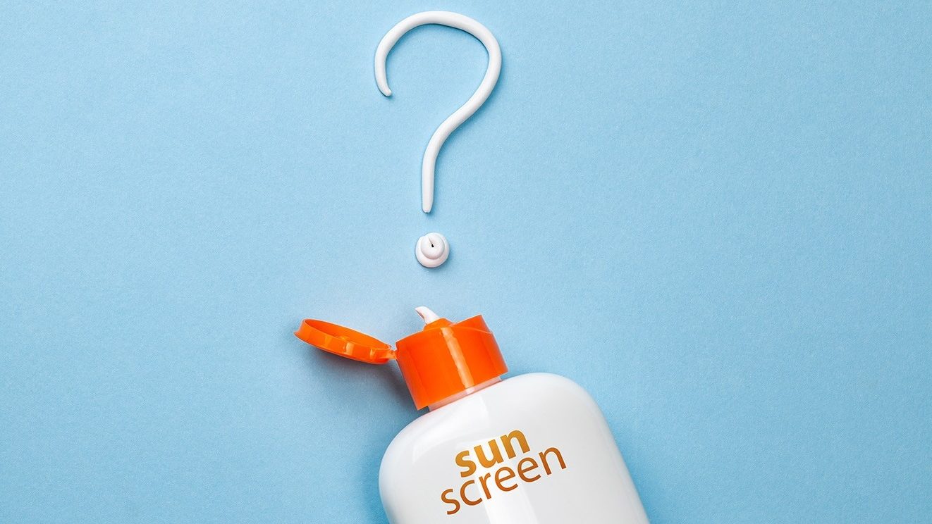 An white sunscreen bottle with an open orange cap and text lies underneath a white question mark made of sunscreen.