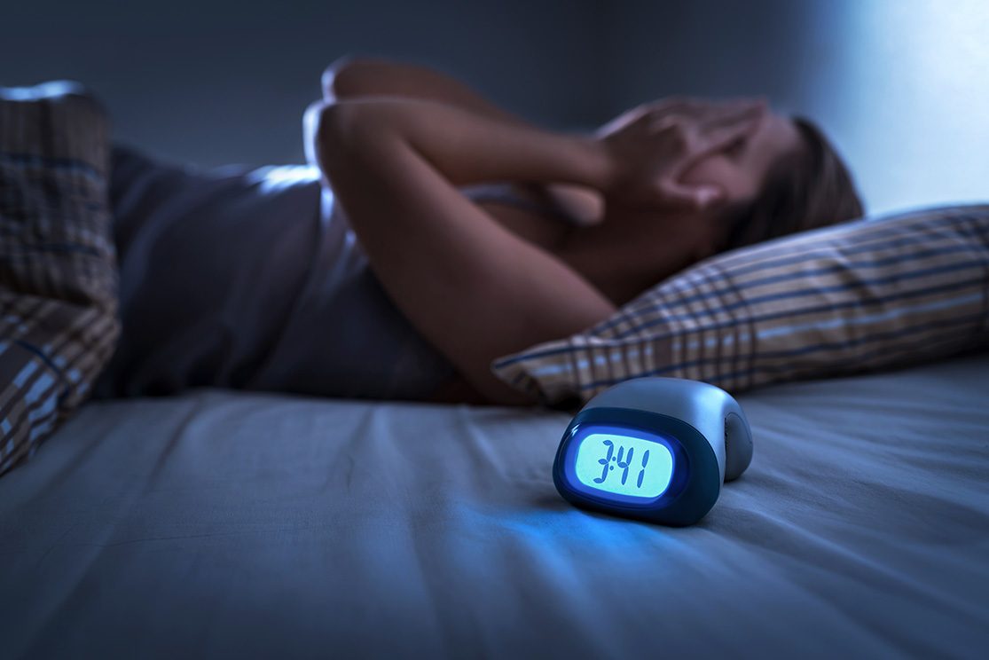 Dimly lit woman in bed covering face in frustration, as nearby digital clock reads 3:41 a.m.