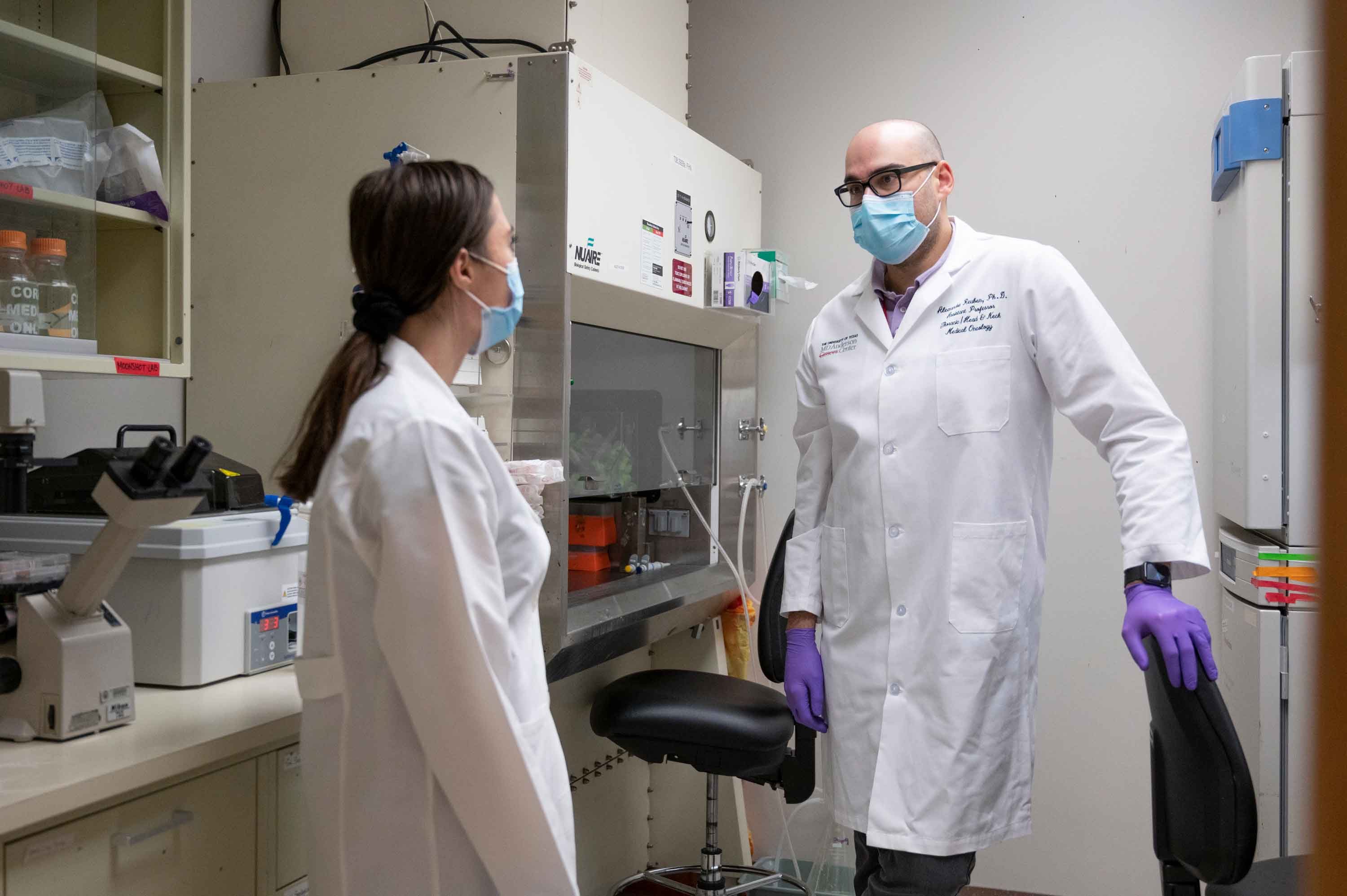 Alexandre Reuben, Ph.D., speaks with a female lab member in the lab. Both are wearing white coats, purple latex gloves and surgical masks.