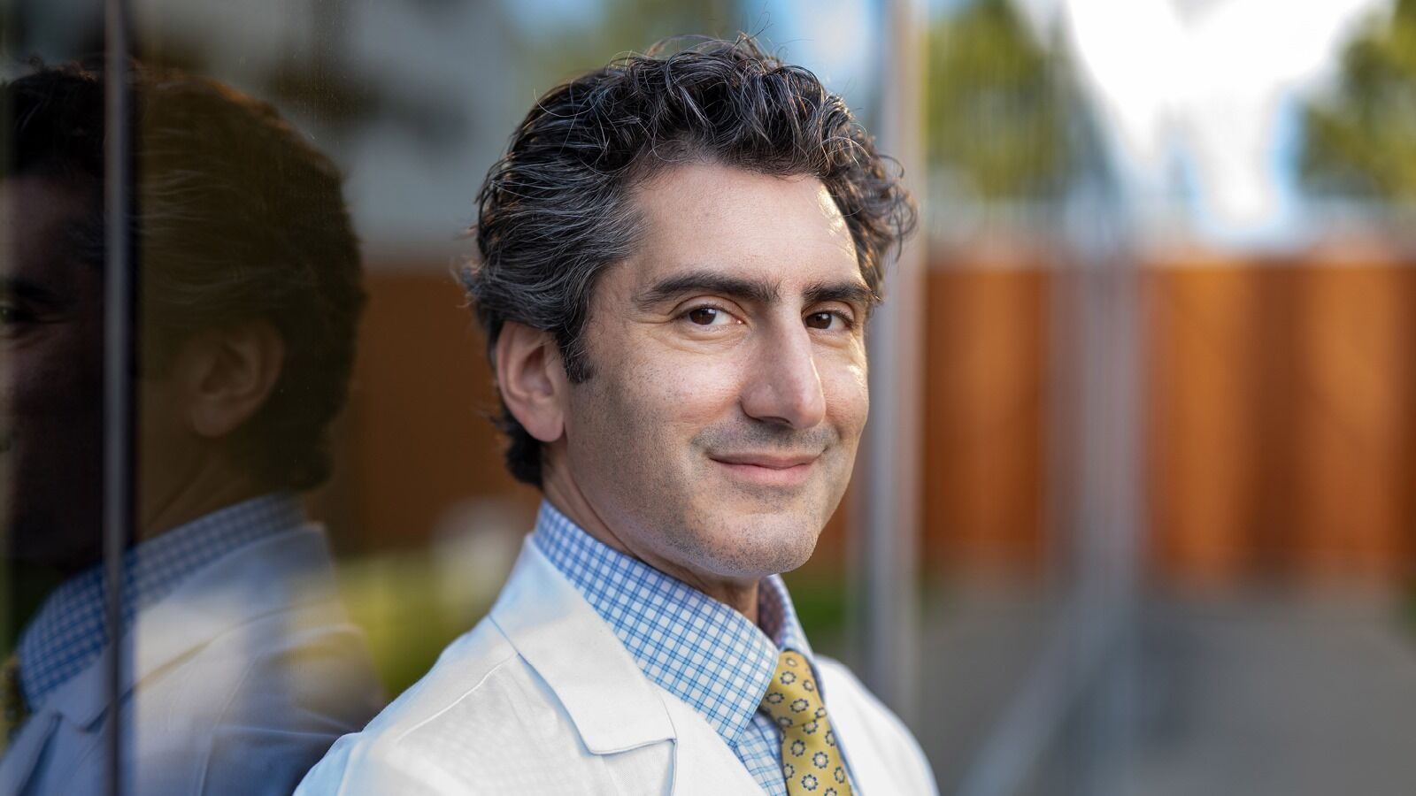 A man in a white coat, blue gingham shirt and yellow tie smiles while standing against a glass wall.