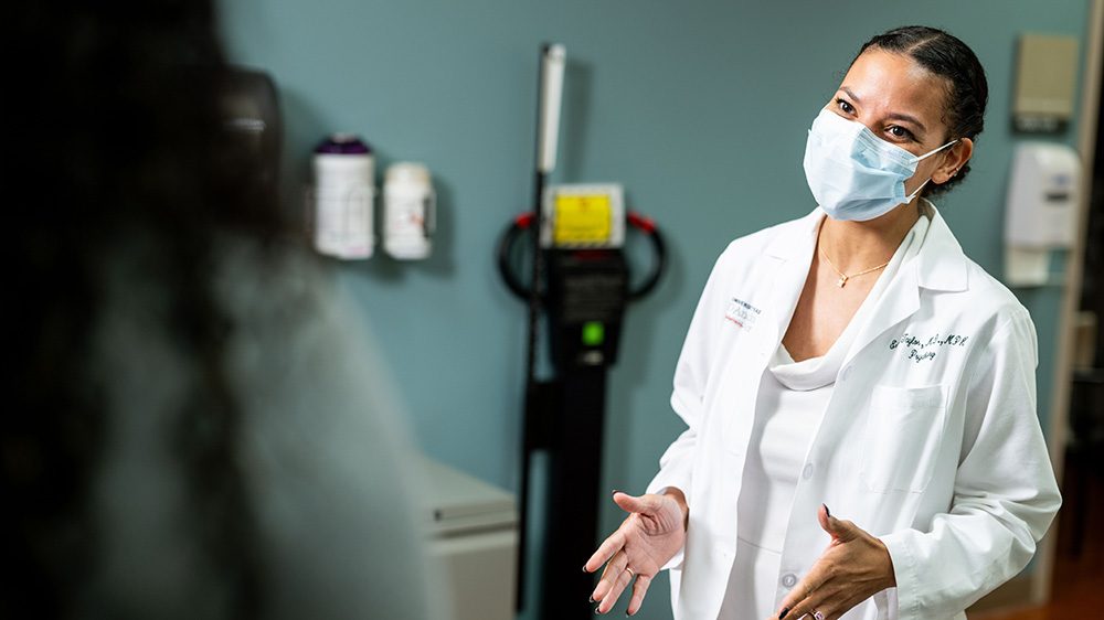 Woman wearing white coat, white blouse and a blue face mask speaks to a person out of frame.